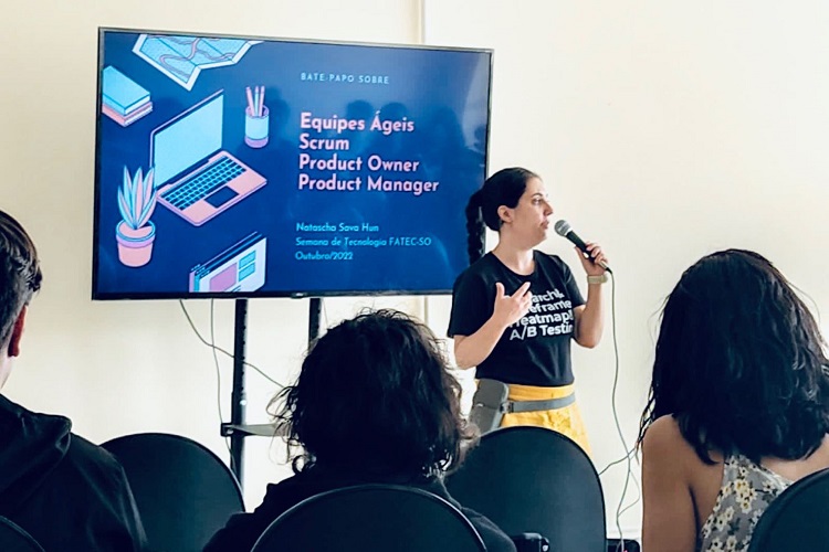 Natascha Hun segurando microfone em palestra sobre equipes ágeis com algumas pessoas assistindo sentadas em cadeiras