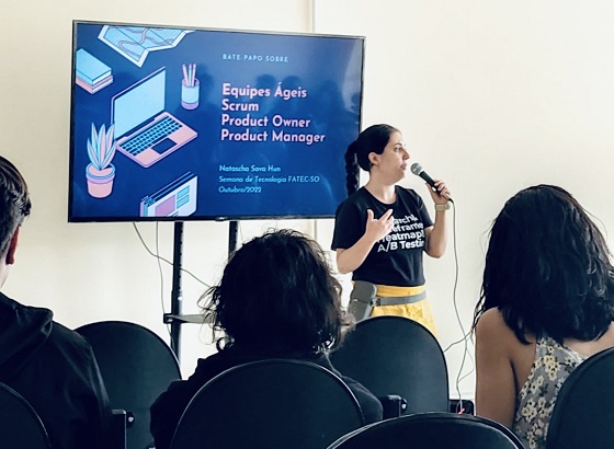 Natascha Hun segurando microfone em palestra sobre equipes ágeis com algumas pessoas assistindo sentadas em cadeiras